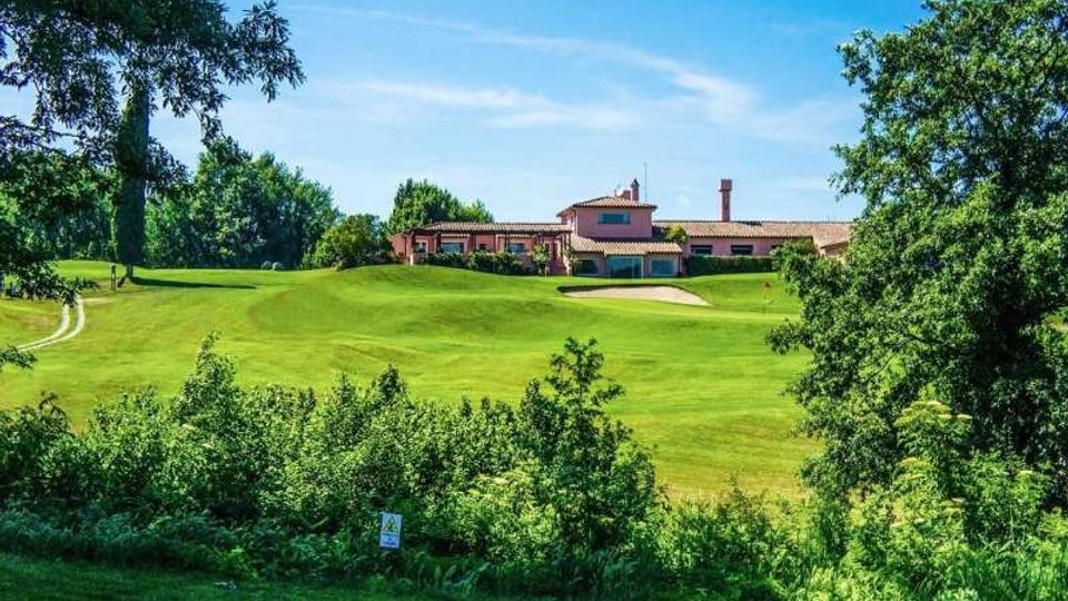 Ampio campo da golf con prati ondulati e un bunker di sabbia, circondato da alberi e vegetazione. Sullo sfondo si vede una club house in stile rustico con tetto in coppi, sotto un cielo azzurro sereno.