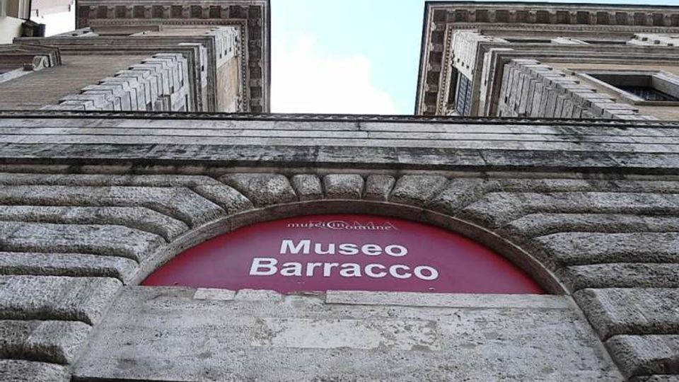 Ingresso del Museo Barracco visto dal basso, con arco in pietra bugnata e insegna rossa “Museo Barracco” sopra il varco. Ai lati si elevano due facciate in muratura con cornici e finestre, incorniciate dal cielo sullo sfondo.