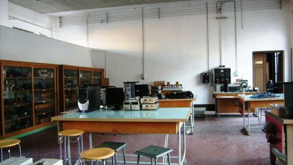 Interno di un laboratorio scolastico di elettronica con banchi da lavoro e sgabelli, strumenti e apparecchiature tecniche appoggiati sui tavoli. Lungo la parete sinistra ci sono armadi vetrati con materiale, soffitto a pannelli e cavi a vista sulle pareti, con una porta sul fondo.