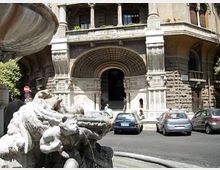 Ingresso di un grande edificio storico in pietra con arco monumentale decorato e colonne, affacciato su una strada urbana. In primo piano si vede una fontana in marmo con figure scolpite e vasca, con auto parcheggiate davanti all’entrata.