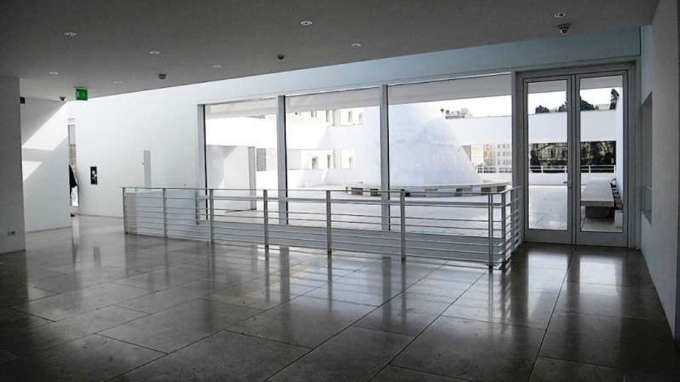 Atrio interno moderno con pavimento in grandi lastre lucide grigie, soffitto con faretti incassati e parapetto in metallo. Ampie vetrate e una porta a vetri si affacciano su una terrazza esterna con elementi architettonici bianchi.