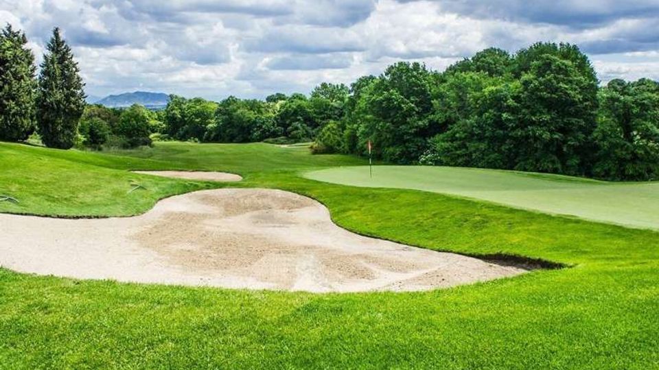 Campo da golf con green e bandierina, circondato da ampi prati e una grande buca di sabbia in primo piano. Sullo sfondo si vedono alberi fitti, colline lontane e un cielo nuvoloso.