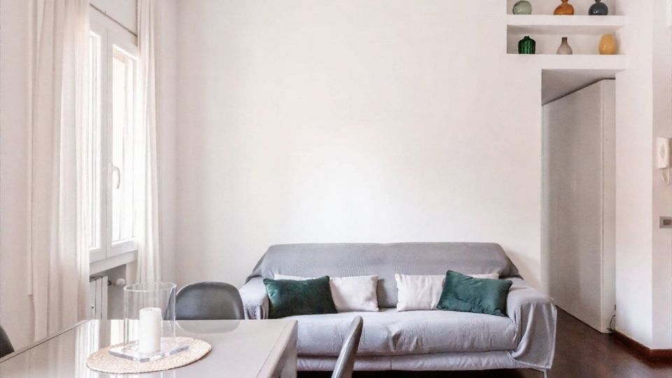 Soggiorno moderno e luminoso con divano grigio e cuscini verdi e chiari, pavimento in legno scuro e tavolo da pranzo con sedie davanti a una grande finestra con tende bianche. A destra si vede un passaggio verso un corridoio e una nicchia a mensole con piccoli vasi decorativi.