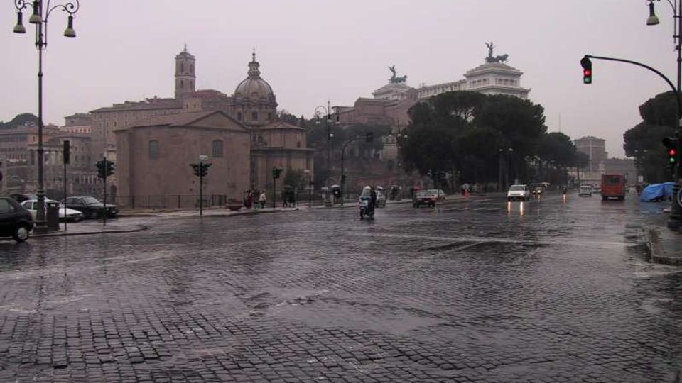 Ampia strada cittadina lastricata in sampietrini bagnati dalla pioggia, con lampioni e semafori lungo i lati e traffico sparso. Sullo sfondo si vedono edifici storici con cupole e campanile, alberi ad alto fusto e un grande monumento su un’altura.