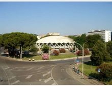 Edificio moderno a pianta circolare con grande cupola bianca e struttura esterna a travi, situato al centro di un’area verde con alberi e aiuole fiorite. In primo piano si vede una rotatoria stradale, con palazzi e vegetazione sullo sfondo.