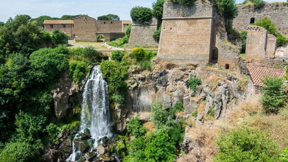 Cascata che scende da una scogliera rocciosa circondata da vegetazione fitta, accanto a mura e bastioni in pietra di una fortificazione storica. Sullo sfondo si vedono edifici in muratura e alberi su un altopiano sopra la gola.