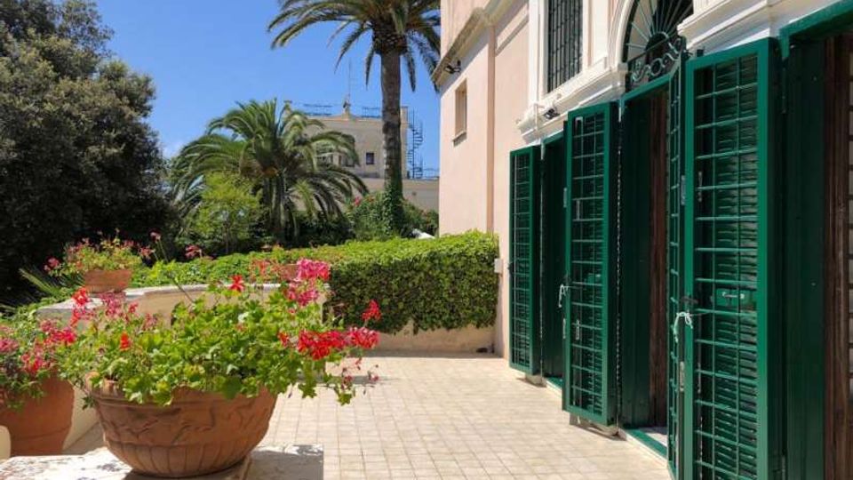 Terrazza esterna di un edificio in stile mediterraneo con facciata chiara e grandi porte-finestre ad arco dotate di persiane verdi aperte. Pavimentazione in piastrelle con fioriere di gerani e giardino con siepi e palme sullo sfondo sotto cielo sereno.
