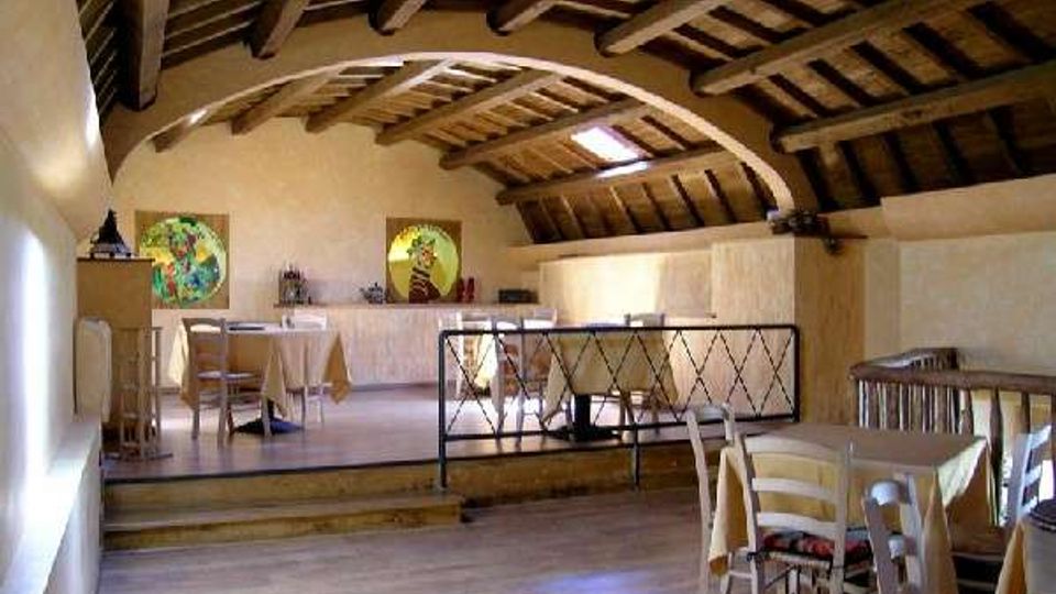 Interno di una sala tipo ristorante con soffitto mansardato in legno e travi a vista, pareti chiare e pavimento in legno. Tavoli apparecchiati e sedie sono disposti su due livelli, separati da un gradino e da una ringhiera metallica, con piccoli lucernari sul tetto.