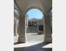 Porticato in pietra con grandi archi che incorniciano una scalinata monumentale in una piazza lastricata. In fondo si vede un edificio storico con loggiato ad arcate, mentre a lato della scalinata sono presenti una fontana e una statua.