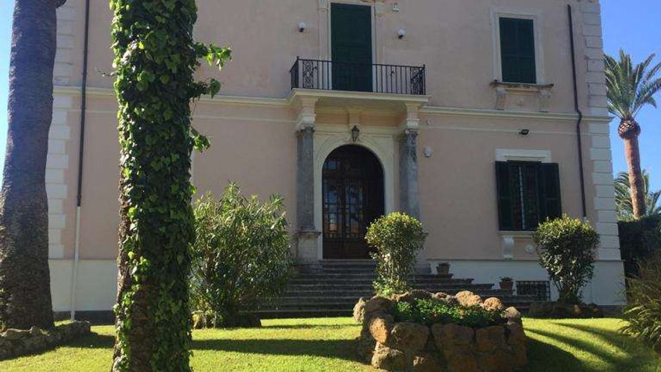 Facciata di una villa storica a tre livelli color beige con ingresso ad arco, balcone centrale e persiane verdi. Davanti si estende un prato curato con palme e aiuole in pietra, sotto un cielo sereno.