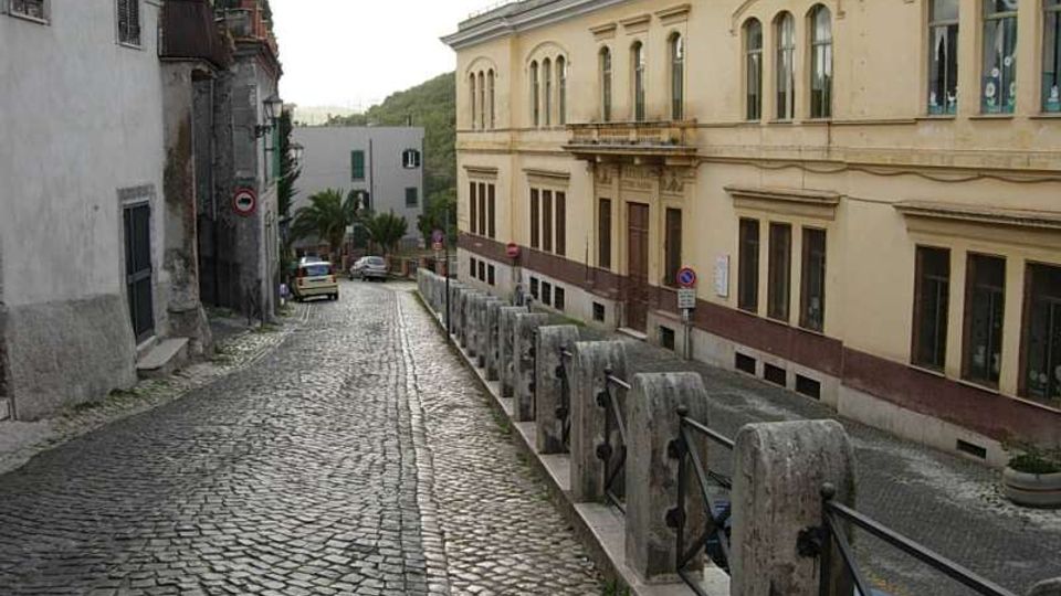 Strada in discesa con pavimentazione in sampietrini, affiancata da edifici storici e da una balaustra in pietra con corrimano metallico. Sulla destra si vede un grande edificio color crema con finestre ad arco, con alcune auto parcheggiate lungo la via.