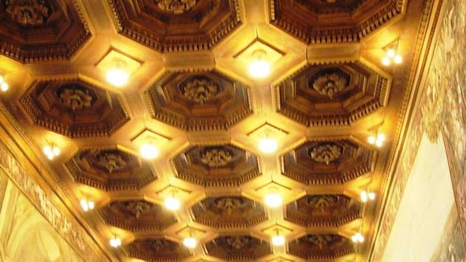 Soffitto a cassettoni ottagonali in legno, illuminato da numerose luci calde, in una lunga sala decorata. Le pareti laterali e di fondo sono rivestite da grandi affreschi con scene figurative e archi dipinti.