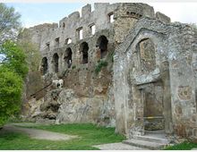 Rovine di un castello in pietra con una lunga fila di archi e aperture, costruite su una parete rocciosa sopra un prato. In primo piano si vede una struttura muraria più bassa con un grande arco e tracce di intonaco, accanto a un sentiero e alberi.