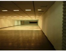 Ampio corridoio sotterraneo con pavimento piastrellato e pareti bianche, illuminato da una griglia di luci rettangolari incassate nel soffitto. La prospettiva conduce verso un’area più buia in fondo, con una breve scalinata e pannelli o vetrate.