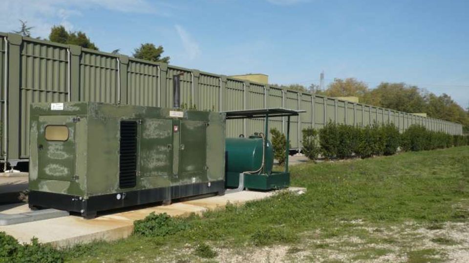 Area industriale all’aperto con un grande generatore verde su una piattaforma di cemento e un serbatoio cilindrico con piccola tettoia. Sullo sfondo si estende una lunga barriera metallica verde con siepi e prato ai lati sotto un cielo sereno.