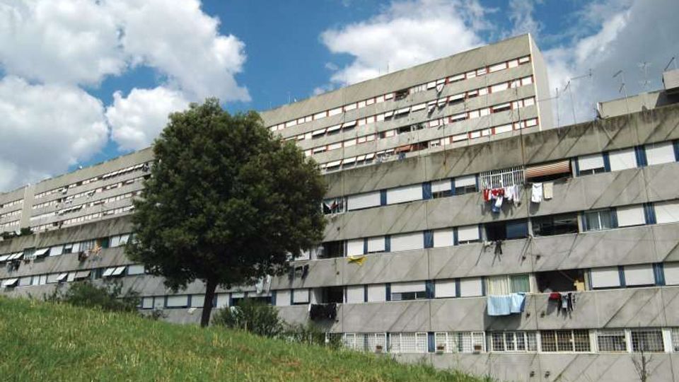 Grande complesso residenziale a più piani in cemento, con lunghe file di finestre e balconi con panni stesi. In primo piano c’è un prato in pendenza con un albero, sullo sfondo cielo azzurro con nuvole.
