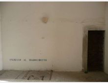 Parete interna intonacata con scritta “INGRESSO AL TRABOCCHETTO” vicino al pavimento e una piccola applique al centro. A destra si vede un’apertura con cornice in pietra che conduce a un vano buio, con pavimento in lastre e un pannello metallico a filo.