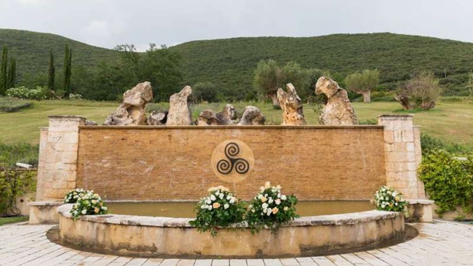 Fontana semicircolare in pietra con vasca bassa e parete in mattoni decorata da un emblema circolare; sopra la parete sporgono grandi rocce ornamentali. Davanti sono disposte fioriere con fiori bianchi e sullo sfondo si vedono colline verdi e alberi in un paesaggio rurale.