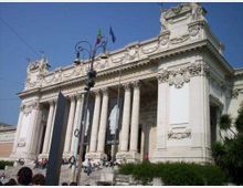 Facciata di un grande edificio monumentale neoclassico con alto portico e colonne corinzie, ricchi bassorilievi e timpano decorato. Davanti si vedono una scalinata, lampioni stradali e bandiere sul tetto, in un contesto urbano.