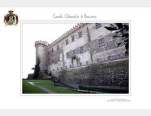 Veduta esterna del Castello Odescalchi di Bracciano, con alte mura in pietra, finestre regolari e una torre cilindrica merlata. In primo piano si vede un prato con un sentiero e il basamento del bastione lungo il perimetro del castello.