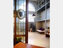Interno di un loft con soffitti alti e grandi finestre ad arco che illuminano uno spazio open space. In primo piano si vede una porta in vetro decorato, mentre sul fondo ci sono un soppalco con ringhiera metallica, una scala, una lampada a sospensione e un divanetto.