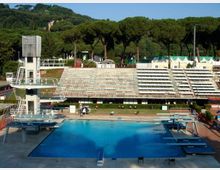 Piscina olimpionica all’aperto con piattaforme e trampolini per tuffi e una torre con scale sul lato sinistro. Sullo sfondo si vedono ampie gradinate in cemento e una fila di pini con una collina boscosa dietro.