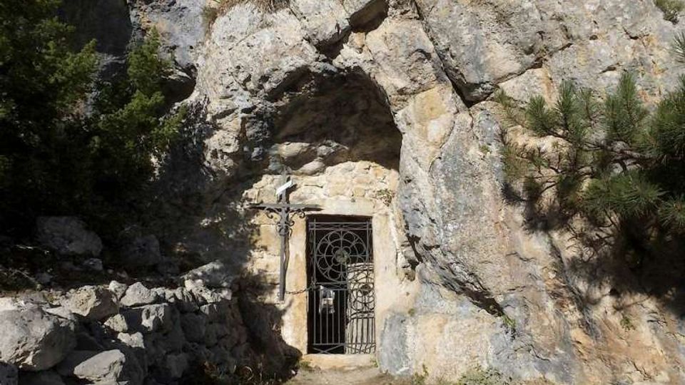 Ingresso di una grotta scavata in una parete rocciosa, con cancello in ferro battuto e una croce fissata alla roccia accanto. L’area circostante è un sentiero sterrato tra massi, vegetazione bassa e pini sulle rocce.
