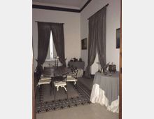Sala da pranzo in stile classico con pareti bianche, alte finestre con tende scure e cornici decorative sul soffitto. Al centro ci sono un tavolo con sedie in ferro battuto su un tappeto e, lungo le pareti, tavoli coperti da tovaglie con oggetti esposti.