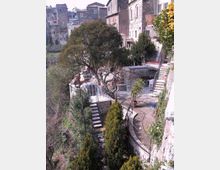 Cortile terrazzato in pietra su un pendio, con scale, parapetti in ferro e un vialetto curvo tra alberi e cespugli. Sullo sfondo si vedono case in pietra addossate l’una all’altra, tipiche di un borgo storico.