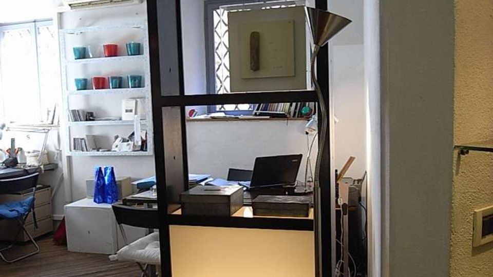 Interno di un ufficio o studio con pavimento in legno e soffitto a volta, separato da una libreria nera con pannelli retroilluminati e oggetti esposti. Sullo sfondo si vedono una scrivania con computer, scaffali bianchi con contenitori colorati e una finestra che illumina l’ambiente.