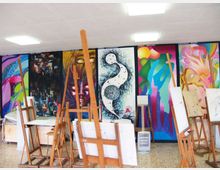 Aula di arte con cavalletti in legno e tele appoggiate, illuminata da pannelli a soffitto. La parete di fondo è coperta da grandi murales colorati e astratti.
