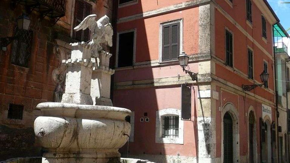 Piazzetta in un centro storico con una fontana in pietra al centro, sormontata da una scultura alata. Intorno si vedono edifici a più piani con facciate in intonaco, finestre con persiane e lampioni a muro lungo una strada stretta.