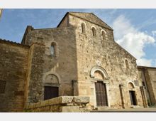 Facciata in pietra di una chiesa romanica con portale centrale ad arco e grandi porte in legno, affiancata da ingressi laterali. La struttura massiccia presenta piccole finestre ad arco in alto ed è ripresa dall’esterno sotto un cielo parzialmente nuvoloso.