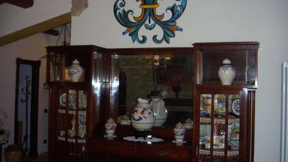 Interno rustico con soffitto a travi in legno e parete bianca decorata da un grande motivo floreale dipinto. Due vetrine in legno scuro con ante in vetro espongono ceramiche e piatti decorati, con un mobile centrale e specchio sullo sfondo.