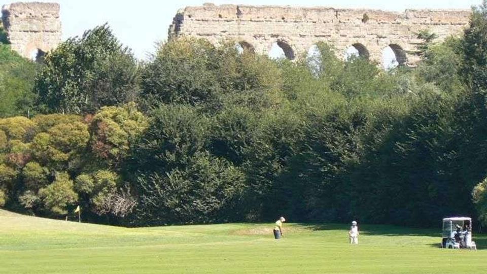 Campo da golf con ampio prato verde in primo piano e alberi fitti sullo sfondo. Dietro la vegetazione si intravedono i resti di un grande acquedotto in pietra con arcate.