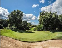 Campo da golf con green circolare e bandierina al centro, circondato da alberi e vegetazione. In primo piano si vede un bunker di sabbia, con cielo azzurro e nuvole sullo sfondo.