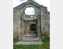 Rovine di un edificio in pietra con facciata a timpano e grande arco centrale, con muratura a vista e intonaco deteriorato. Un passaggio ad arco conduce all’interno, accessibile tramite gradini in pietra, con prato e cielo sullo sfondo.