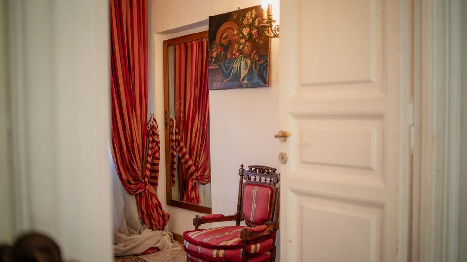Angolo interno in stile classico con poltrona in legno imbottita a righe rosse, specchio alto con tende a righe rosse e tappeto. Sulla parete bianca sono visibili un quadro e una lampada a parete, con una porta bianca in primo piano.