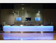 Banco reception moderno con lunga scrivania illuminata da una fascia blu, in una hall con pavimento lucido e pareti scure. Sullo sfondo ci sono schermi informativi e lampade a sospensione sopra il bancone.