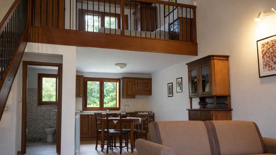 Interno di un appartamento a pianta aperta con soppalco in legno e ringhiera in metallo, scala laterale e soffitto con travi a vista. Al piano inferiore ci sono cucina e zona pranzo con mobili in legno e grandi finestre sul verde, un bagno visibile dalla porta a sinistra e un soggiorno con divano e credenza.