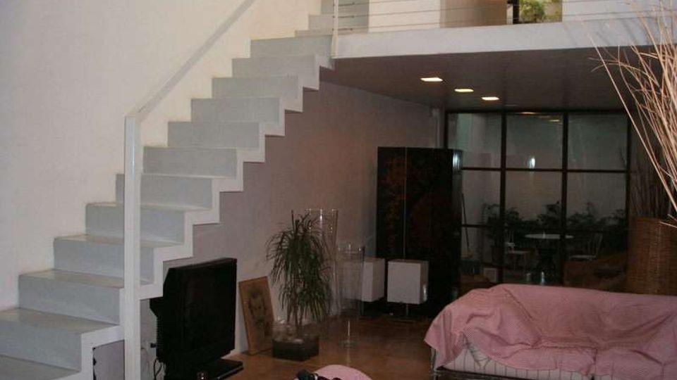 Interno di un loft moderno con scala bianca a vista che conduce a un soppalco con ringhiera metallica. Soggiorno open space con divano basso, TV, pavimento in legno e parete vetrata sul fondo che separa un’area con piante e arredi.
