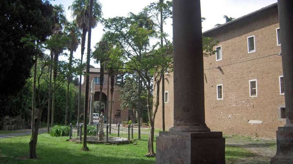 Cortile erboso con alberi e palme, affiancato da un lungo edificio in mattoni con finestre rettangolari. In primo piano si vede una grande colonna di portico e, sullo sfondo, un arco in laterizio che conduce a un passaggio.
