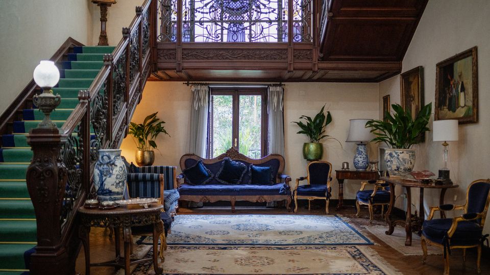 Interno di un salone in stile classico con grande scala in legno e ringhiera in ferro battuto, soppalco con vetrata decorata e pavimento in parquet con ampi tappeti. Divani e poltrone blu, tavolini con lampade e vasi, piante in vaso e quadri alle pareti vicino a una portafinestra con tende.