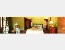 Camera da letto con pareti verde chiaro, letto matrimoniale al centro con testiera decorata, pavimento in cotto e mobili in legno. A sinistra c’è una finestra con tende a motivo floreale e una sedia in vimini, mentre a destra un comò con lampada e un’apertura verso un bagno.