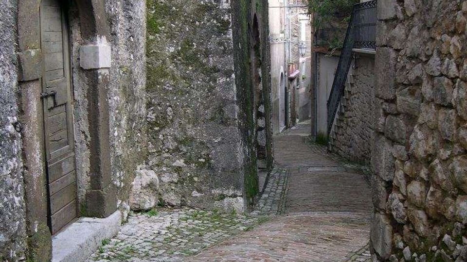 Vicolo stretto in un borgo storico con pavimentazione in ciottoli e mattoni, in lieve discesa tra edifici in pietra e intonaco segnati dal tempo. Si vedono una porta ad arco, un lampione a parete e muri ravvicinati che incanalano la strada.