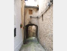 Vicolo stretto in pietra tra edifici in muratura, con pavimentazione irregolare e un piccolo arco che collega le pareti sopra il passaggio. Sulle facciate si vedono cavi e tubazioni a vista, una finestra piccola e alcuni vasi appesi.