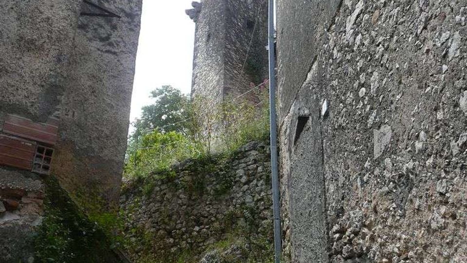 Vicolo stretto in un borgo antico con scalini in pietra invasi da erba e vegetazione, affiancato da alte pareti di case in pietra grezza e intonaco. Sullo sfondo si vedono muri a secco e un edificio con tetto sporgente e tubazioni esterne.