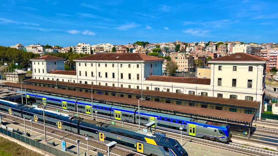 Vista panoramica di una stazione ferroviaria con più binari e treni blu in sosta sotto pensiline lunghe. Sullo sfondo si vede un grande edificio storico chiaro con tetti in tegole rosse e, oltre, un quartiere collinare con palazzi.