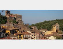 Panorama di un borgo collinare con case addossate dai tetti in coppi e facciate colorate, dominato da una torre cilindrica e da un grande edificio storico in pietra. A destra si vede un campanile e, sullo sfondo, un’area boscosa con colline sotto un cielo sereno.