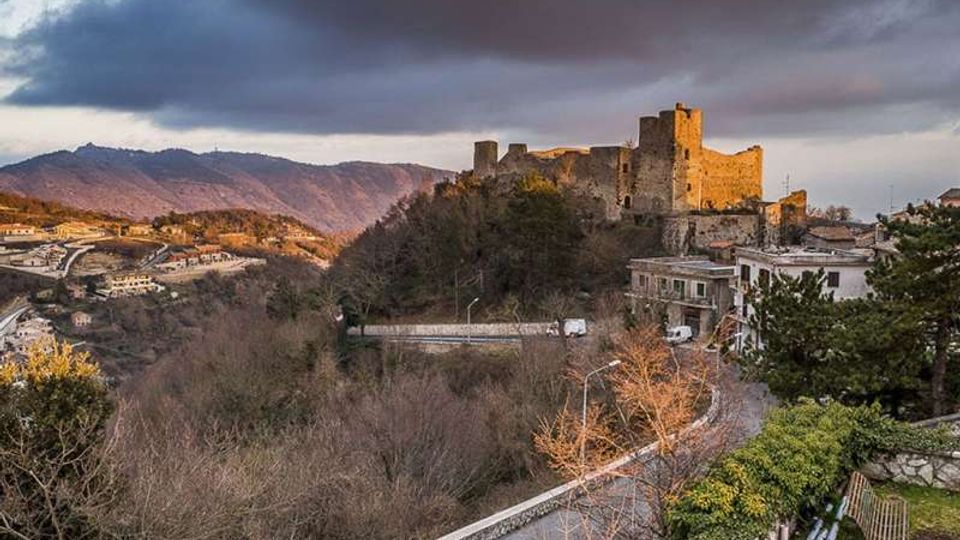 Castello in pietra su un’altura, con torri e mura illuminate, che domina una valle boscosa. In primo piano una strada curva con muretto e alcune case ai margini del pendio, sullo sfondo montagne sotto un cielo nuvoloso.